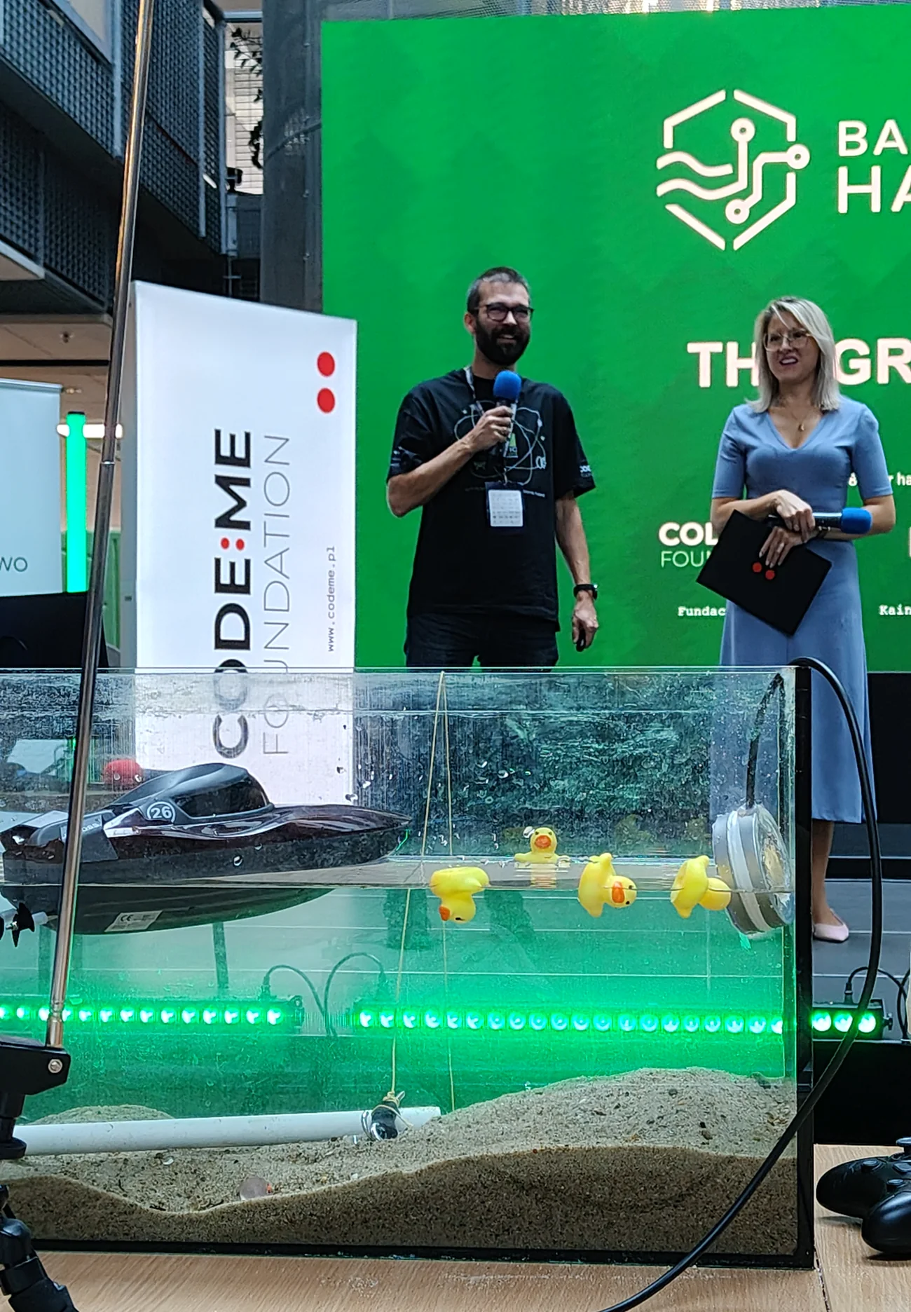 Akwarium z piaskiem, zakopaną rurą, czujnikiem, motorówką i kaczuszkami, w tle scena hackathonu