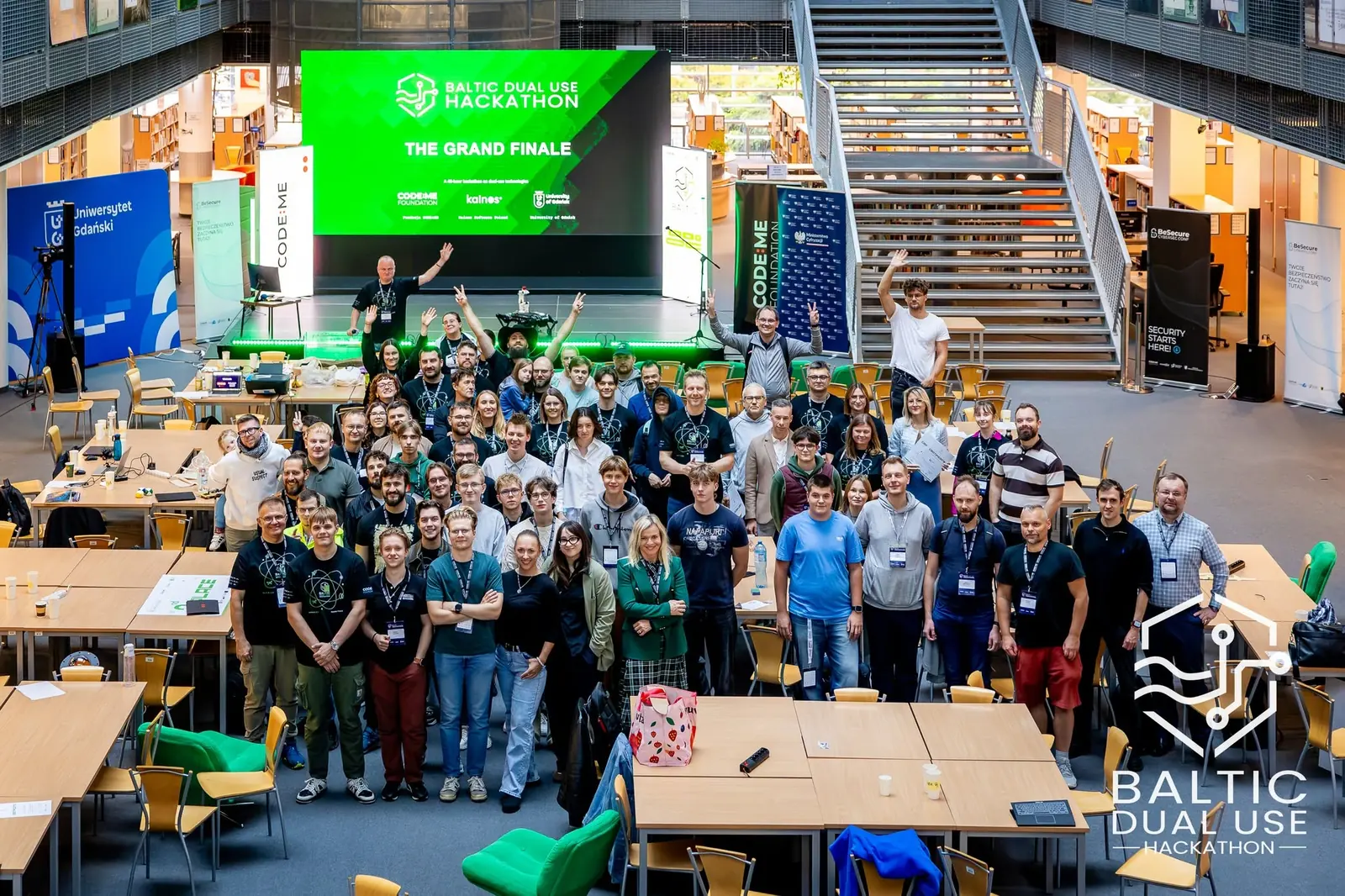 Wszyscy uczestnicy Baltic Dual Use Hackathon przed sceną z napisem The Grand Finale w hali biblioteki
