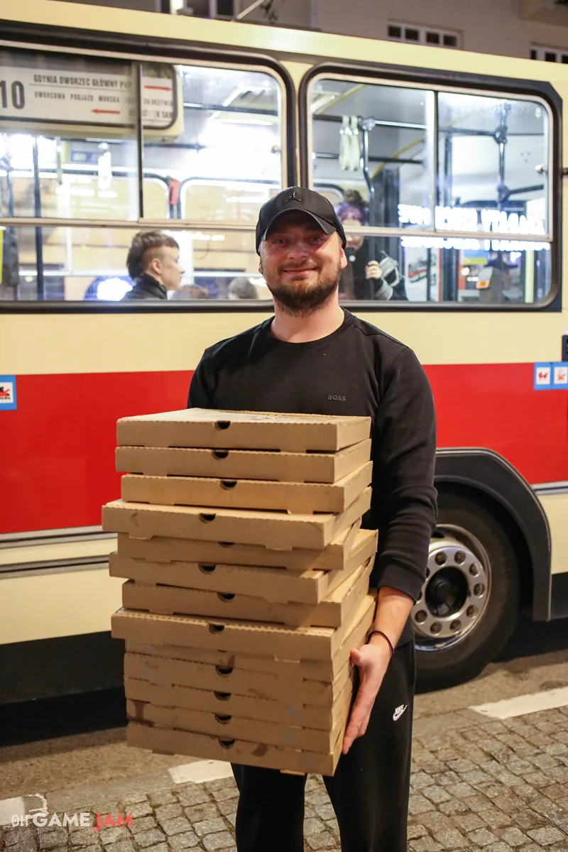 Uśmiechnięty mężczyzna niesie stos pudełek z pizzą obok trolejbusu