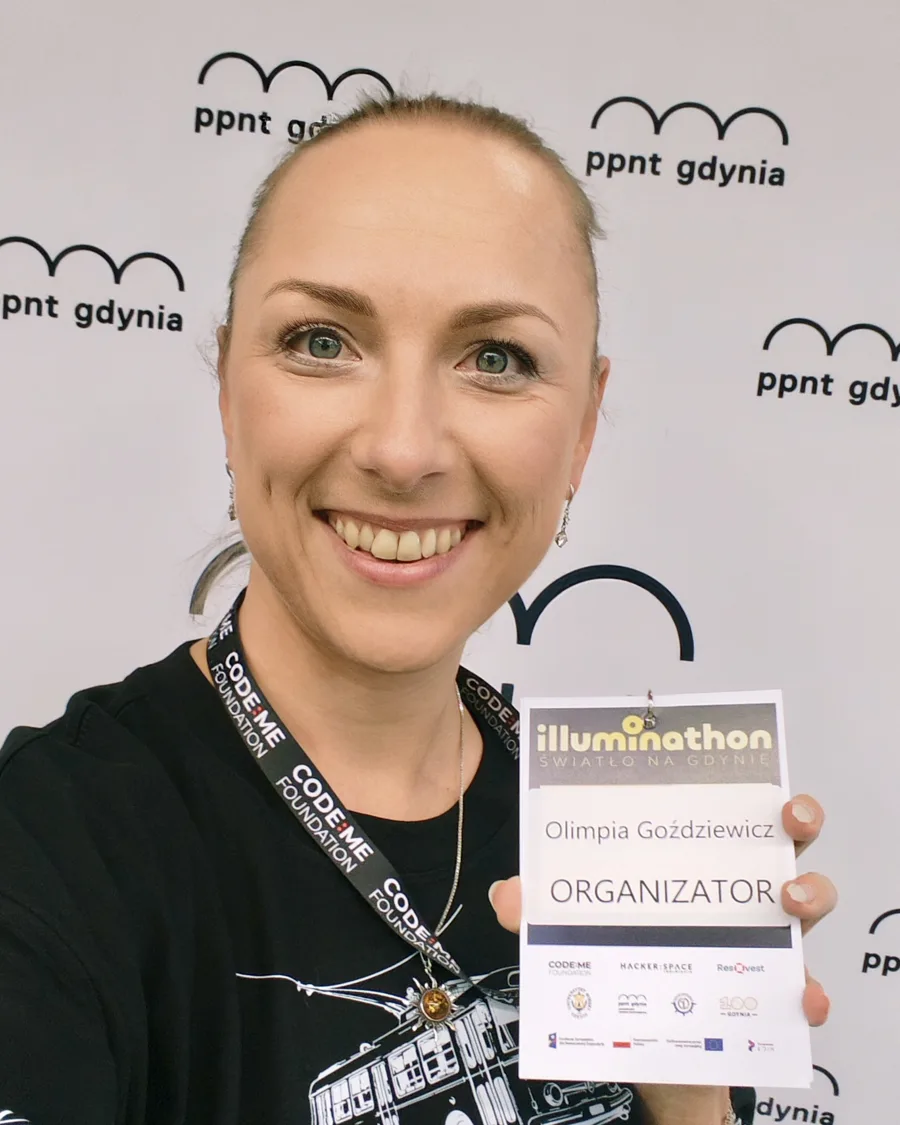 Olimpia Goździewicz na tle ścianki PPNT Gdynia, w koszulce Illuminathonu, pokazuje identyfikator z napisem Organizator