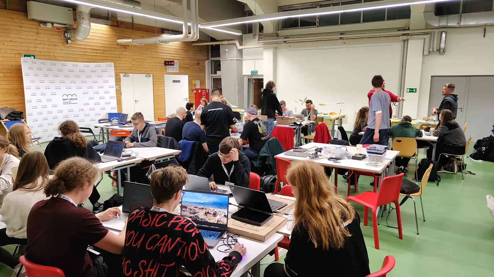 Pełna sala w piątek wieczorem: zespoły przy stołach z laptopami i elektroniką, w tle ścianka PPNT Gdynia