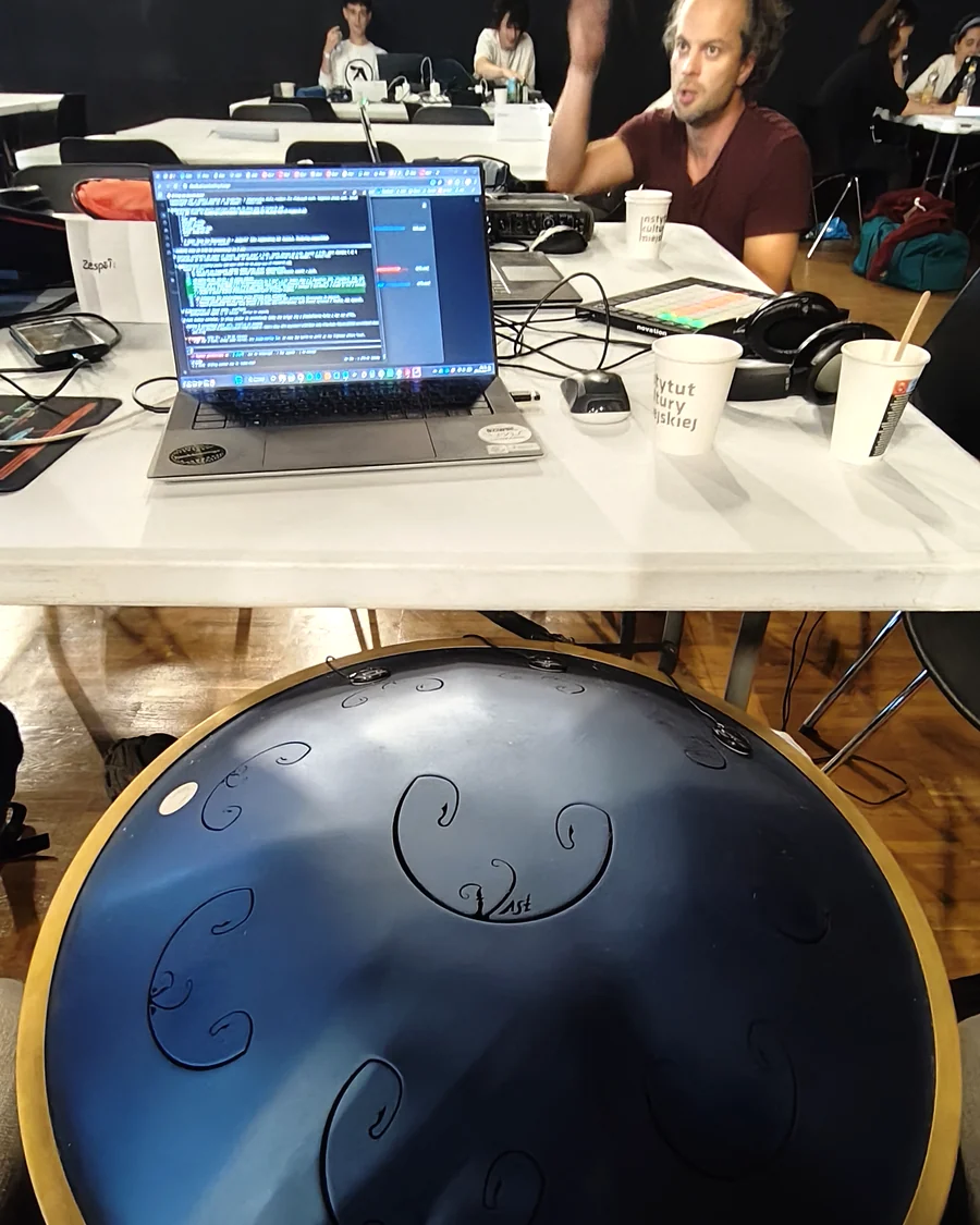Handpan oparty o kolana, w tle stół z laptopem i pracujący uczestnicy